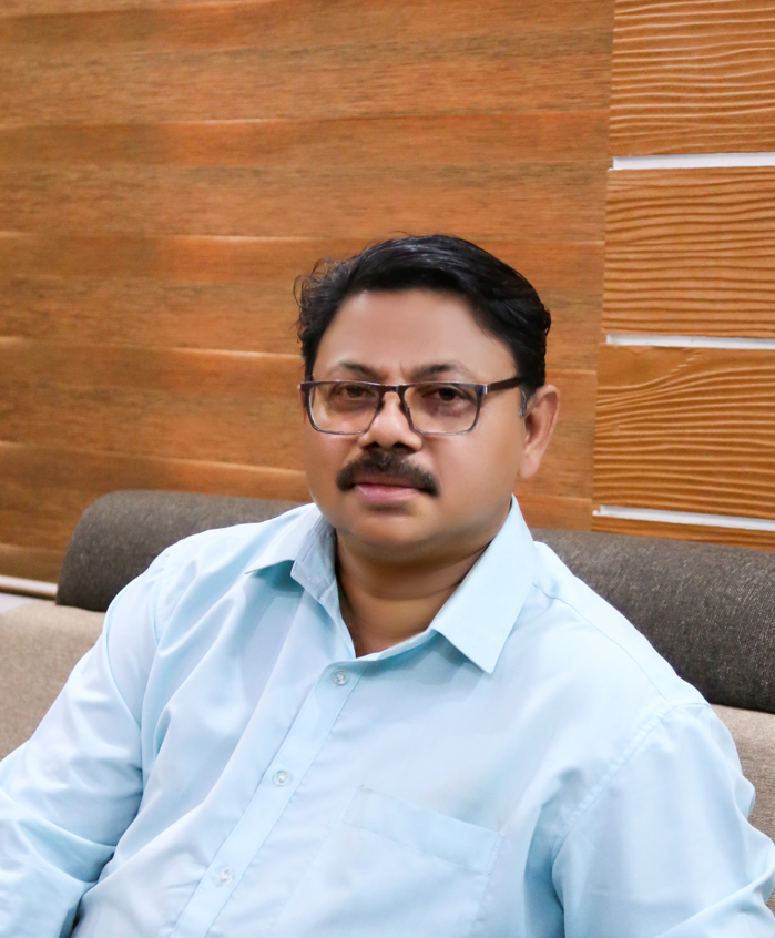 A professional-looking man with dark hair and a beard, sitting in an office setting, wearing a dark suit and blue shirt. He is posing confidently with a smile, and his hand rests near his chin. A wooden background and a desk with a blue laptop are visible.Yembroos Animal Feeds