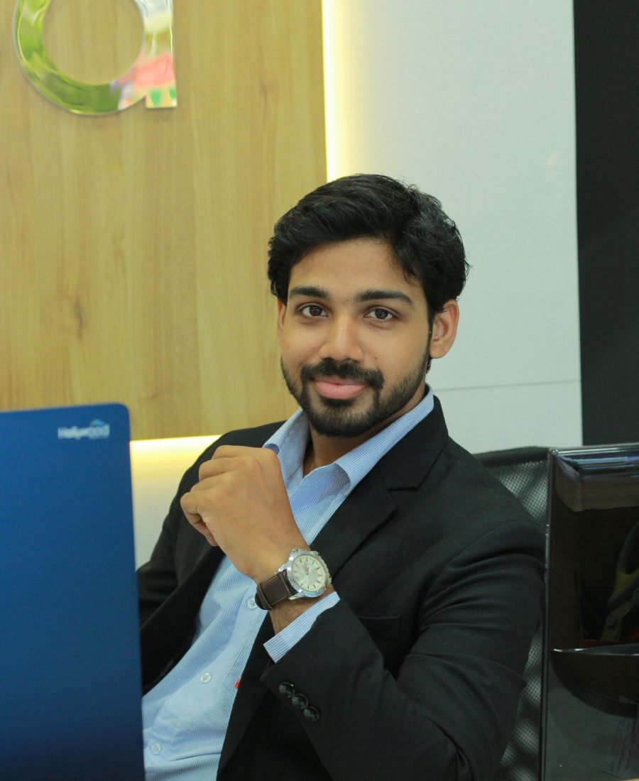 A professional-looking man with dark hair and a beard, sitting in an office setting, wearing a dark suit and blue shirt. He is posing confidently with a smile, and his hand rests near his chin. A wooden background and a desk with a blue laptop are visible.Yembroos Animal Feeds