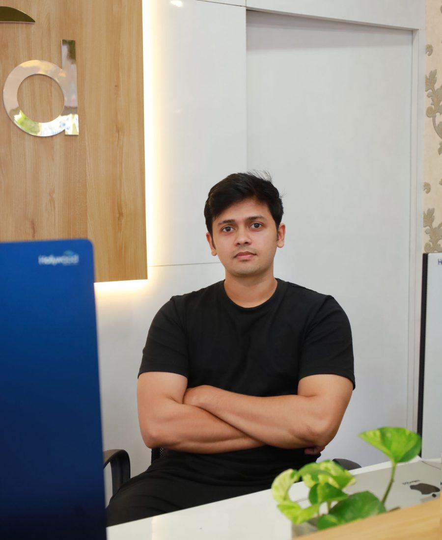 A professional-looking man with dark hair and a beard, sitting in an office setting, wearing a dark suit and blue shirt. He is posing confidently with a smile, and his hand rests near his chin. A wooden background and a desk with a blue laptop are visible.Yembroos Animal Feeds